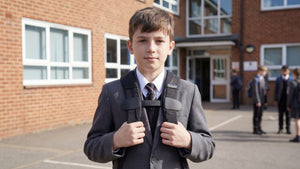 Young boy wearing a KLiPP in a school uniform standing outside a school building with other students in the background.